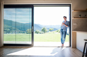 Home front view of young woman with coffee standing by patio door at home.